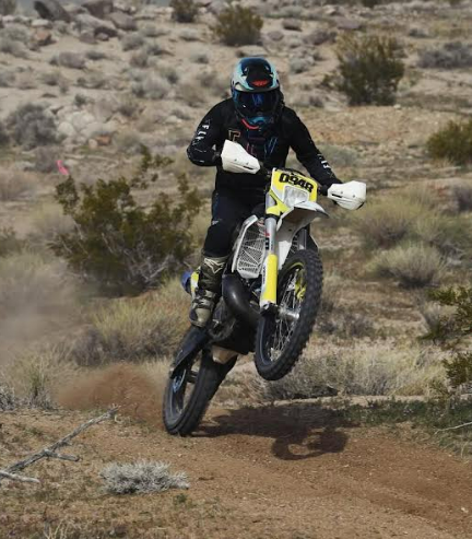Dirt biker in black gear pops a wheelie on a desert trail, with scrub bushes and rocky terrain.