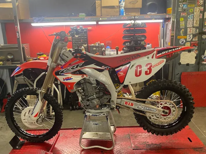 Red and white motocross dirt bike on a stand in a garage, with tools and shelves in the background